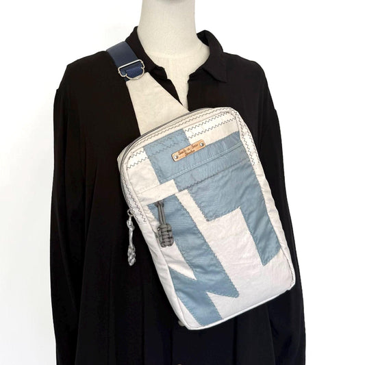 May include: A crossbody sling bag featuring a rectangular shape with a white and dusty blue color-blocked design. The bag has a dark blue adjustable strap, a front zipper closure with a grey cord pull, and a small rectangular metal brand tag.