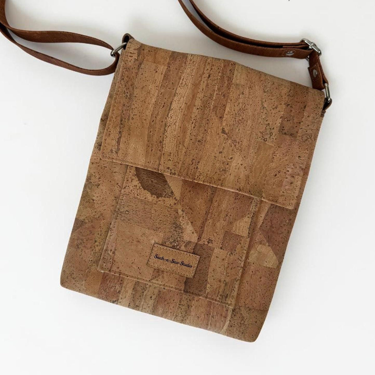 May include: A rectangular crossbody bag crafted from natural tan cork material with a distinct speckled texture. The bag features a fold-over flap, a front pocket with a small rectangular label reading "Cork and Sea Studio," and a brown strap.