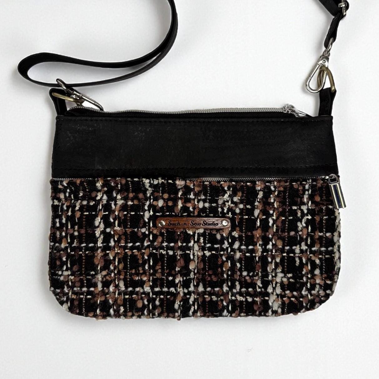 May include: A black crossbody bag with a tweed pattern in brown, white, and black. The bag has a black leather top and strap, with silver-tone hardware. A small, rectangular tag is attached to the front.