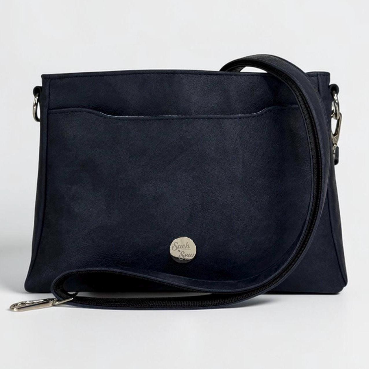 May include: A navy blue crossbody bag with a front pocket and a long, adjustable strap. The bag features a silver-colored clasp and a small, round emblem on the front. The bag is made of a textured material.