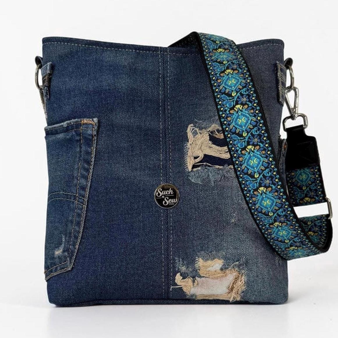 May include: A dark blue denim crossbody bag featuring distressed patches, a side pocket, and a detachable shoulder strap with a vibrant teal, yellow, and black floral pattern. A small circular logo patch with the text Such Sew is centered.