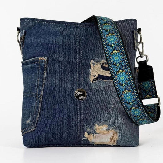 May include: A dark blue denim crossbody bag featuring distressed patches, a side pocket, and a detachable shoulder strap with a vibrant teal, yellow, and black floral pattern. A small circular logo patch with the text Such Sew is centered.
